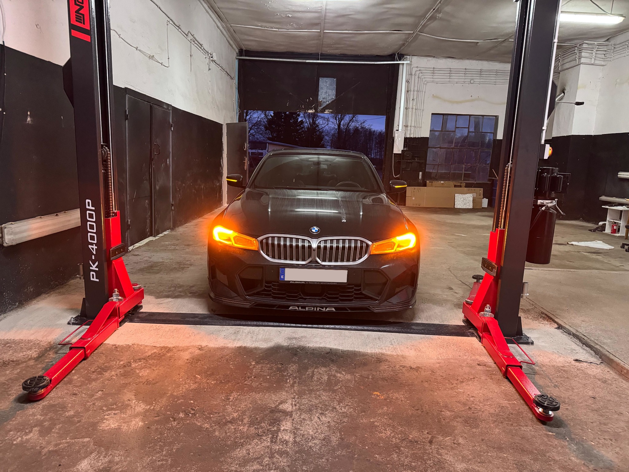 Czarny samochód marki Alpina na podnośniku dwukolumnowym w warsztacie. Włączone światła mijania. Widoczne narzędzia i wyposażenie warsztatowe w tle.