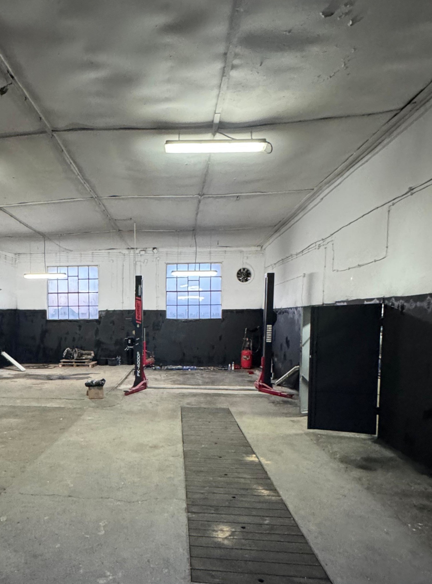 Wnętrze warsztatu samochodowego z podnośnikami i oknami. Widoczne narzędzia i elementy wyposażenia. Surowy, industrialny charakter przestrzeni.