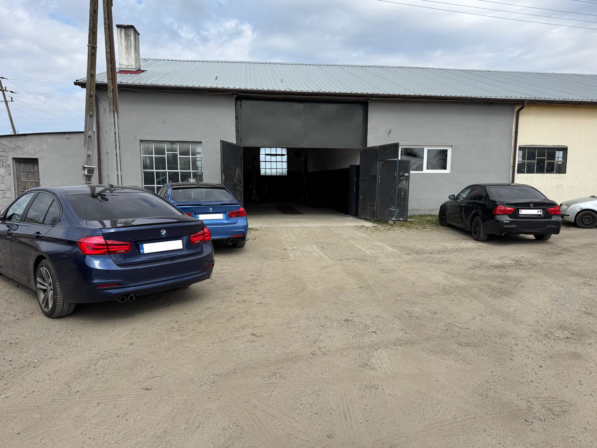 Warsztat samochodowy z otwartą bramą i zaparkowanymi trzema BMW serii 3 w kolorach: granatowym, niebieskim i czarnym na żwirowym placu.