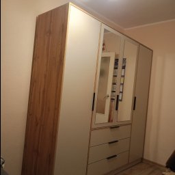 Kamil Nowicki - Czterodrzwiowa szafa z lustrami i szufladami, z drewnianymi bokami, stoi na panelach podłogowych w pokoju o beżowych ścianach. Widoczne oświetlenie sufitowe.