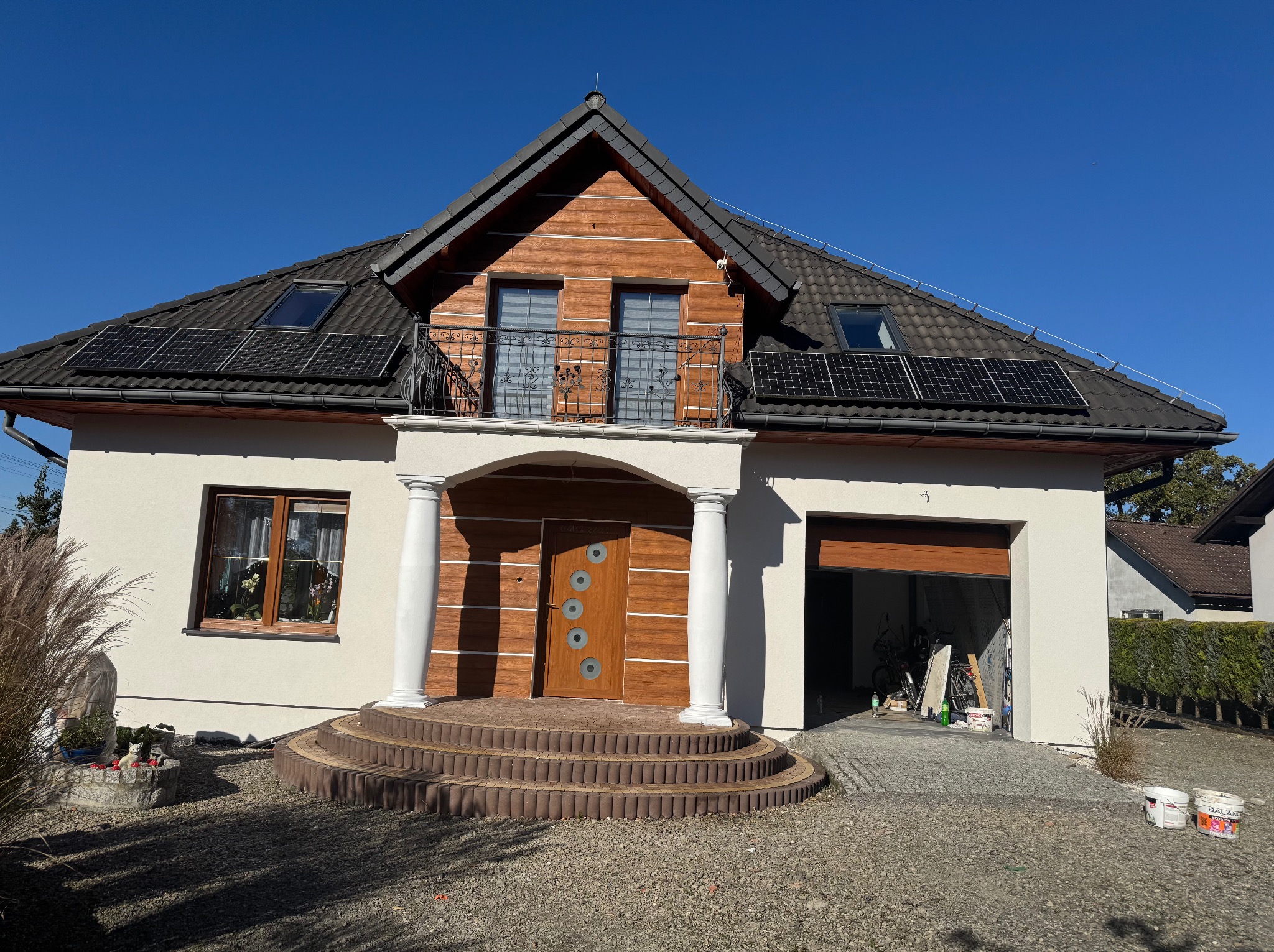 Dwukondygnacyjny dom z jasną elewacją, drewnianą górą i balkonem z kutą balustradą. Na dachu panele słoneczne. Otwarty garaż z widocznym wyposażeniem.