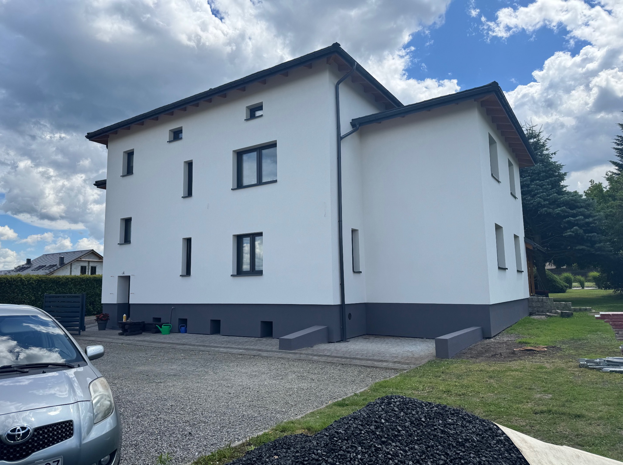 Dwukondygnacyjny dom z białą elewacją i szarym cokołem, widoczny z boku, z fragmentem samochodu i stertą węgla na pierwszym planie, na tle pochmurnego nieba.