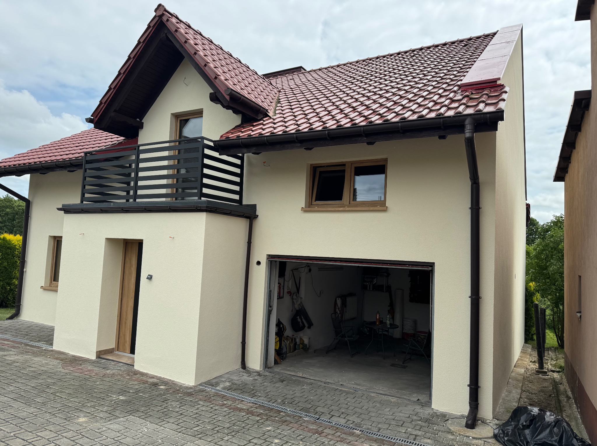 Dwukondygnacyjny dom z beżową elewacją, balkonem z czarną balustradą i otwartym garażem. Czerwona dachówka i kostka brukowa na podjeździe.