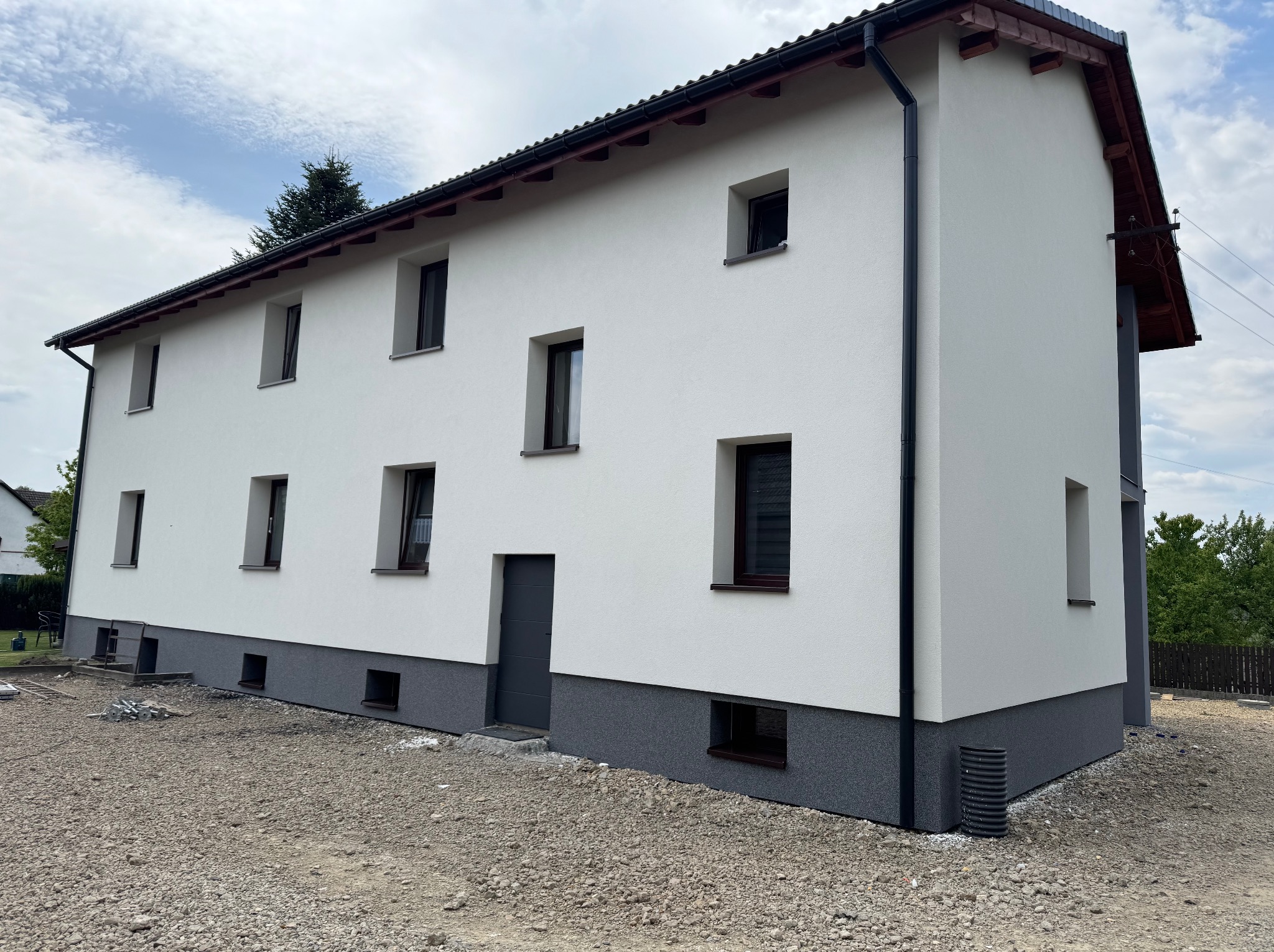 Dwukondygnacyjny dom z białą elewacją i szarym cokołem, widok z zewnątrz. Nowe okna z brązowymi ramami. Teren wokół domu przygotowany pod zagospodarowanie.