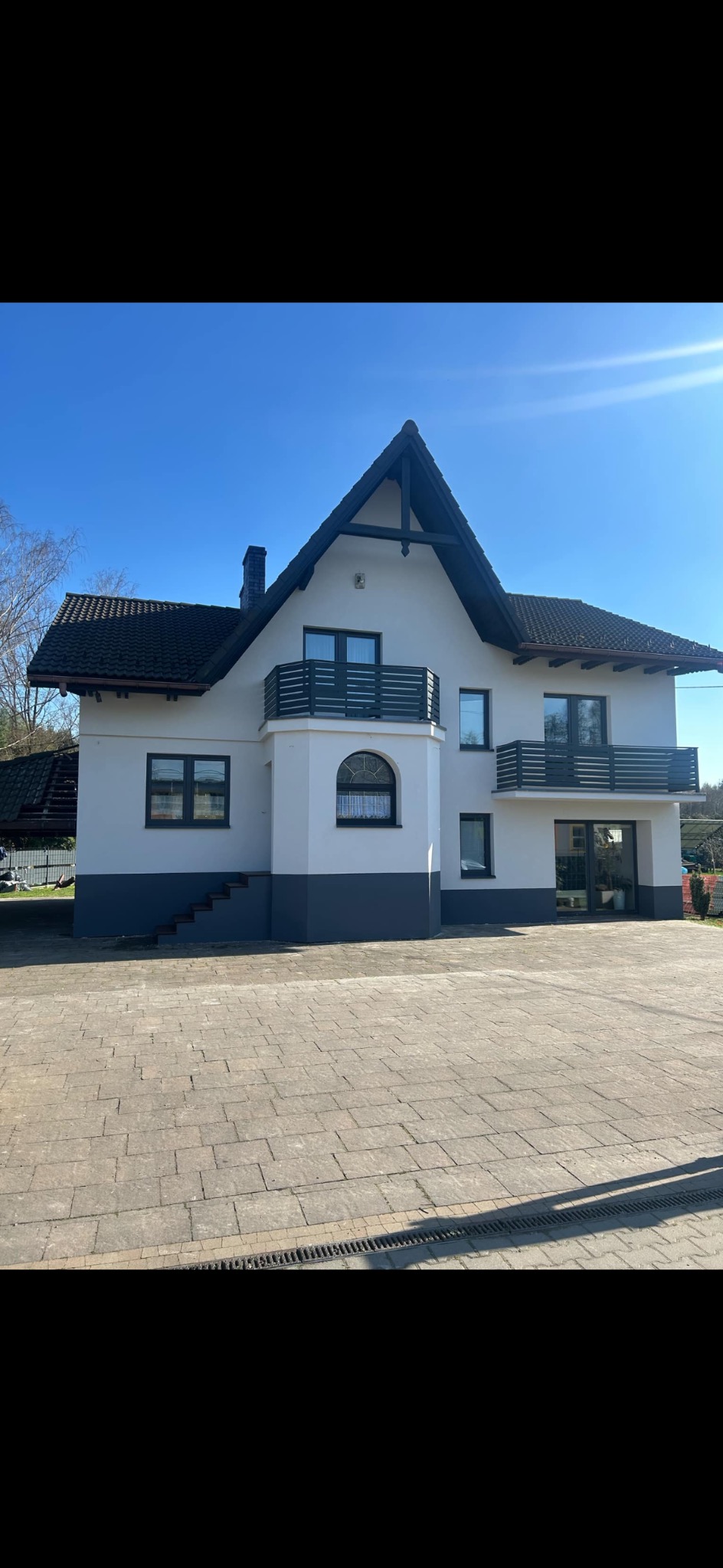 Dwukondygnacyjny dom z balkonami, po remoncie elewacji, z szarą podmurówką i białymi ścianami, widoczny z poziomu brukowanego podjazdu w słoneczny dzień.