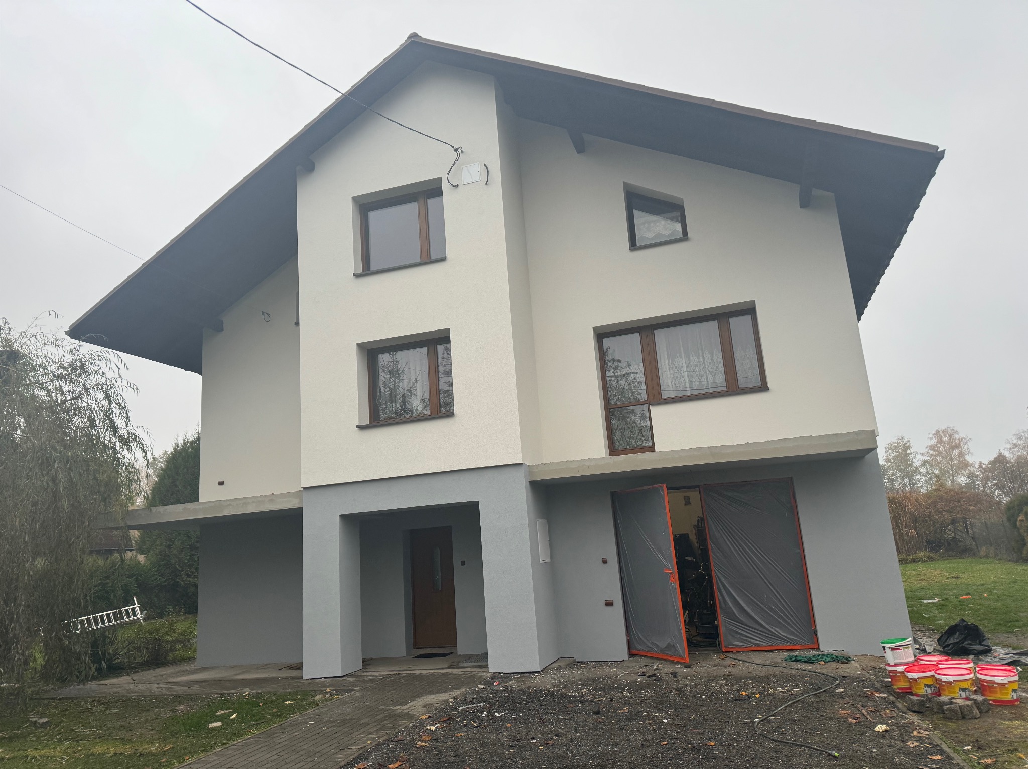 Dwukondygnacyjny dom z jasną elewacją i szarym cokołem. Widoczne otwarte drzwi garażowe zabezpieczone folią i wiaderka z farbą. Pochmurne niebo w tle.