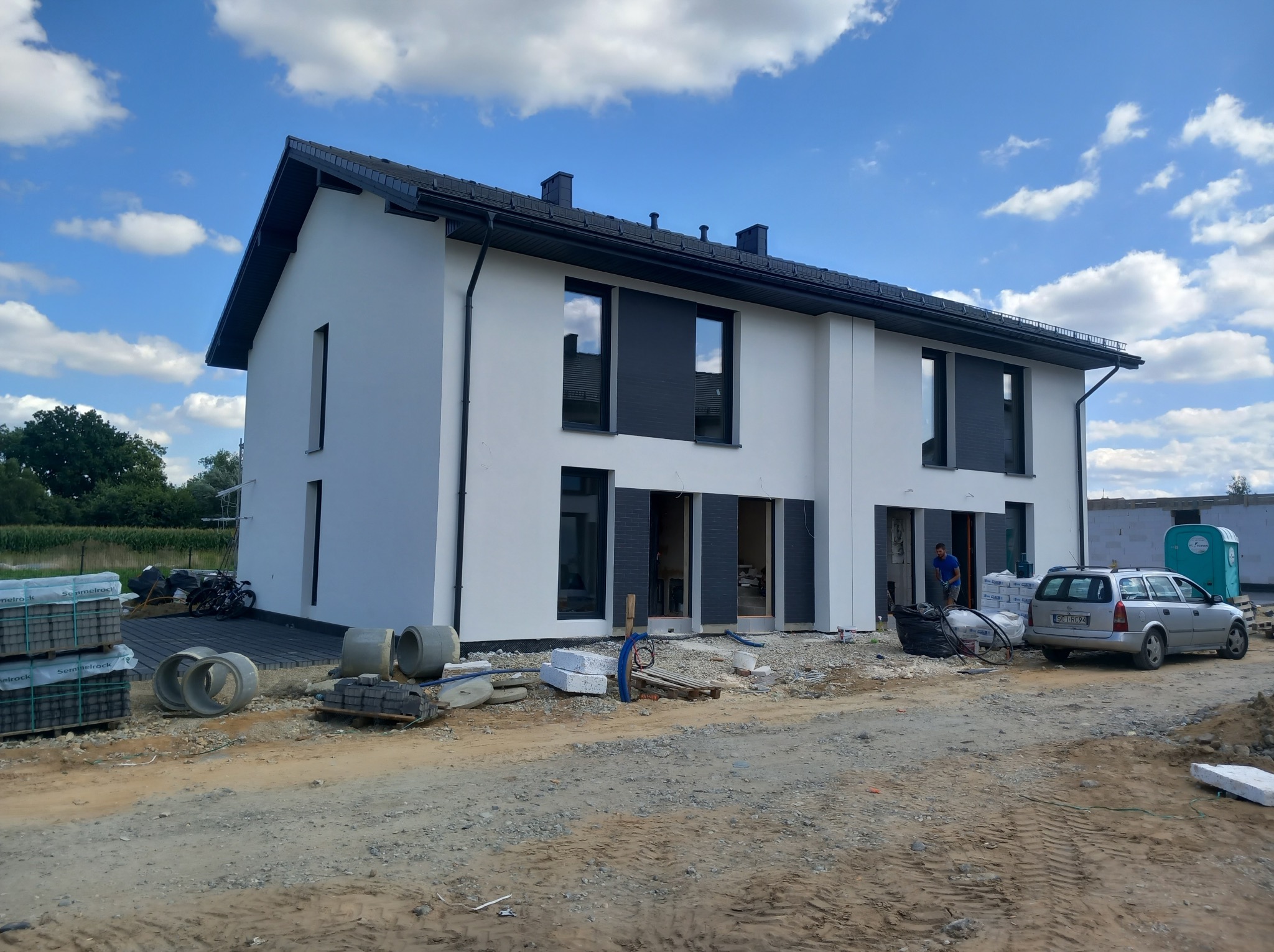 Nowoczesny, dwukondygnacyjny dom z białą elewacją i grafitowymi detalami, w trakcie prac wykończeniowych. Widoczne materiały budowlane i osoba pracująca wewnątrz.