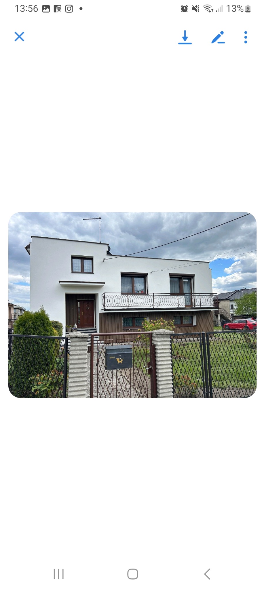 Dwupiętrowy dom z białą elewacją, brązowymi oknami i balkonem z ozdobną balustradą. Widoczny fragment ogrodzenia i skrzynka pocztowa. Pochmurne niebo w tle.