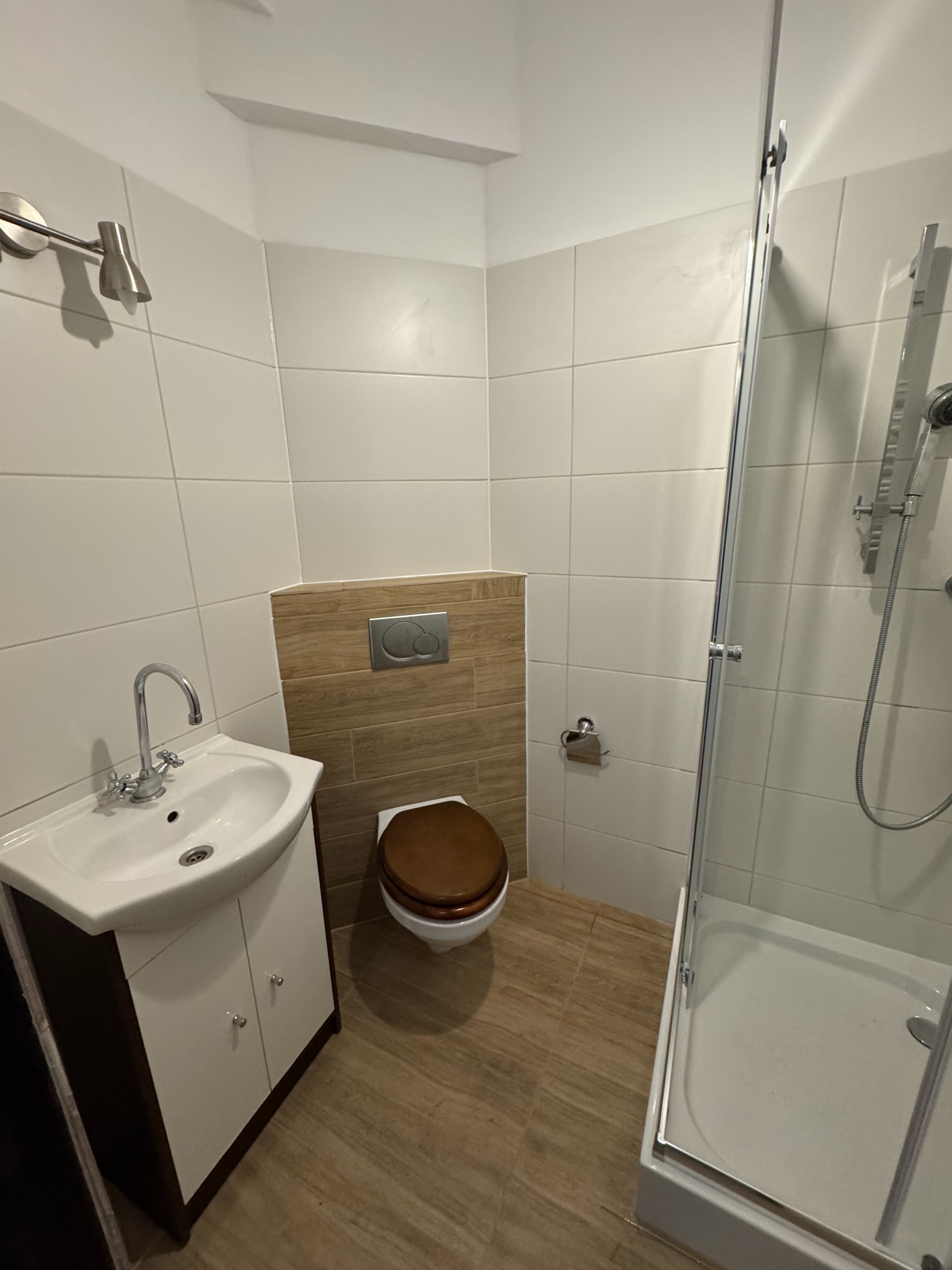 Wykończona łazienka z kabiną prysznicową, umywalką z szafką i toaletą. Jasne płytki na ścianach, drewnopodobne na podłodze i za toaletą. Nowoczesny design.