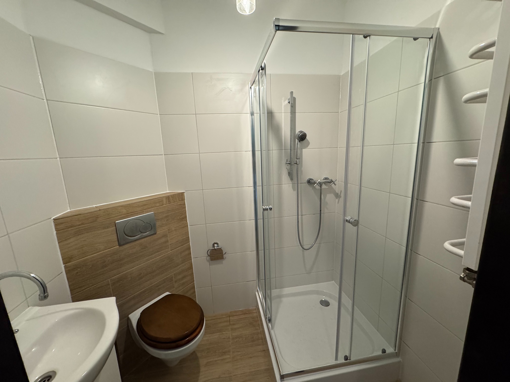 Ukończona łazienka z kabiną prysznicową, umywalką i toaletą. Ściany wyłożone białymi i drewnopodobnymi płytkami. Widoczne detale wykończenia.
