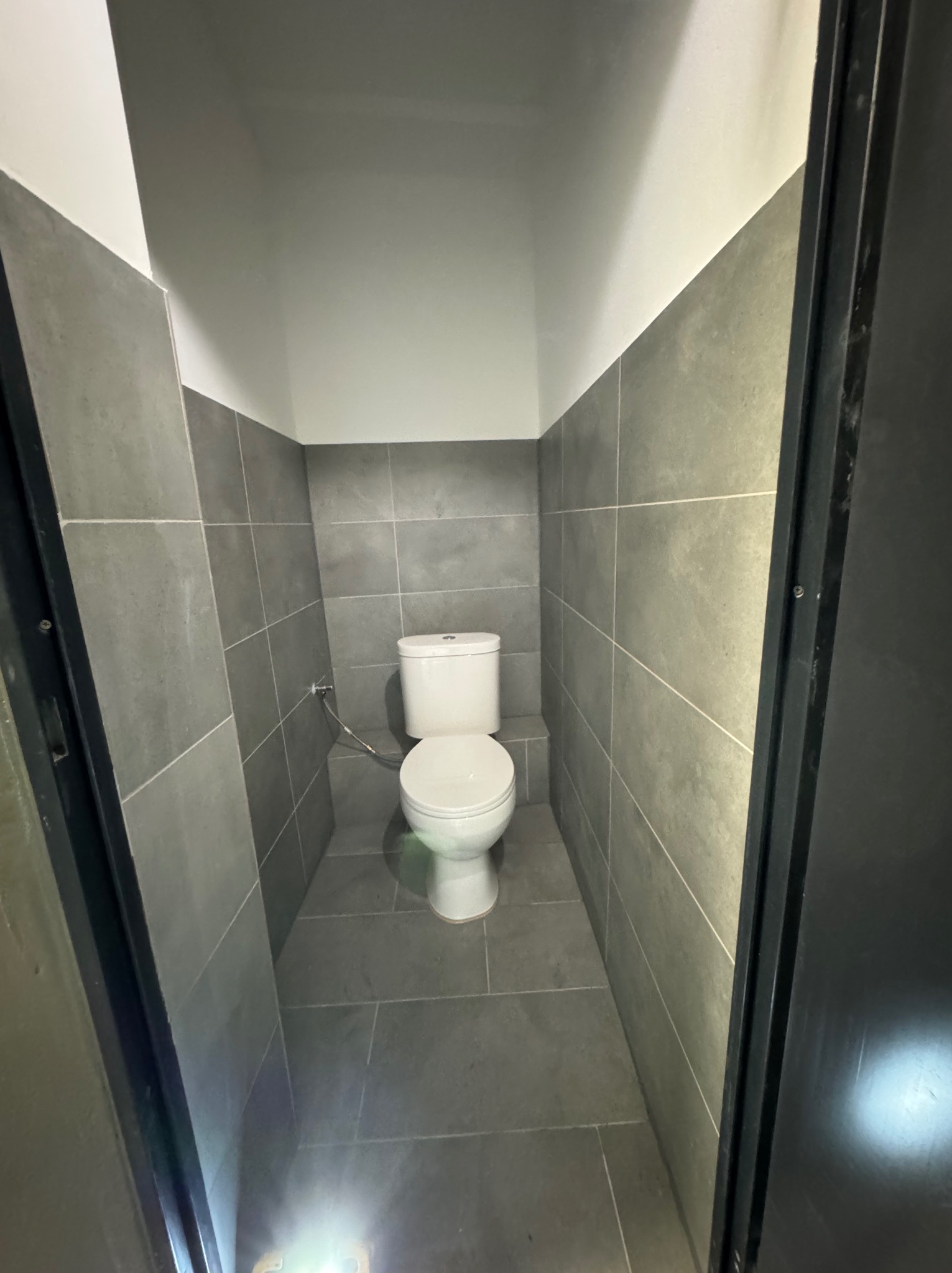 Wykończona łazienka z szarymi płytkami, białą armaturą i czarnymi drzwiami. Minimalistyczny design, widok zza otwartych drzwi. Świeże, nowoczesne wnętrze.