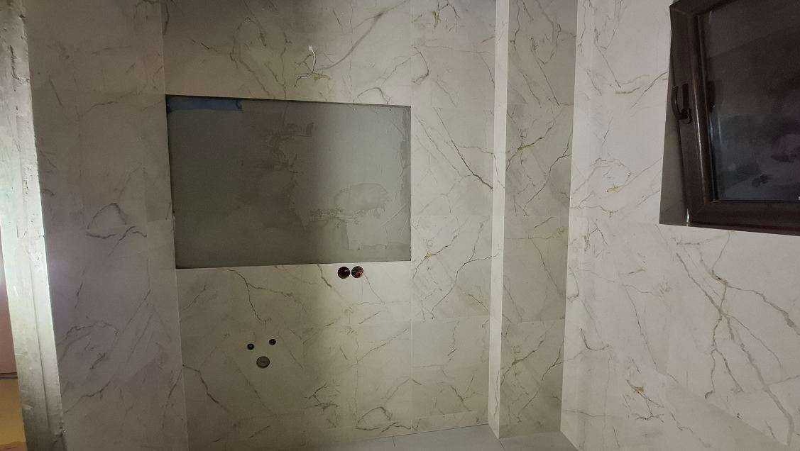 Wykończona ściana łazienki z marmurowymi płytkami, wnęką na lustro i widocznymi punktami instalacji elektrycznej i wodnej. Okno z ciemną ramą w prawym górnym rogu.