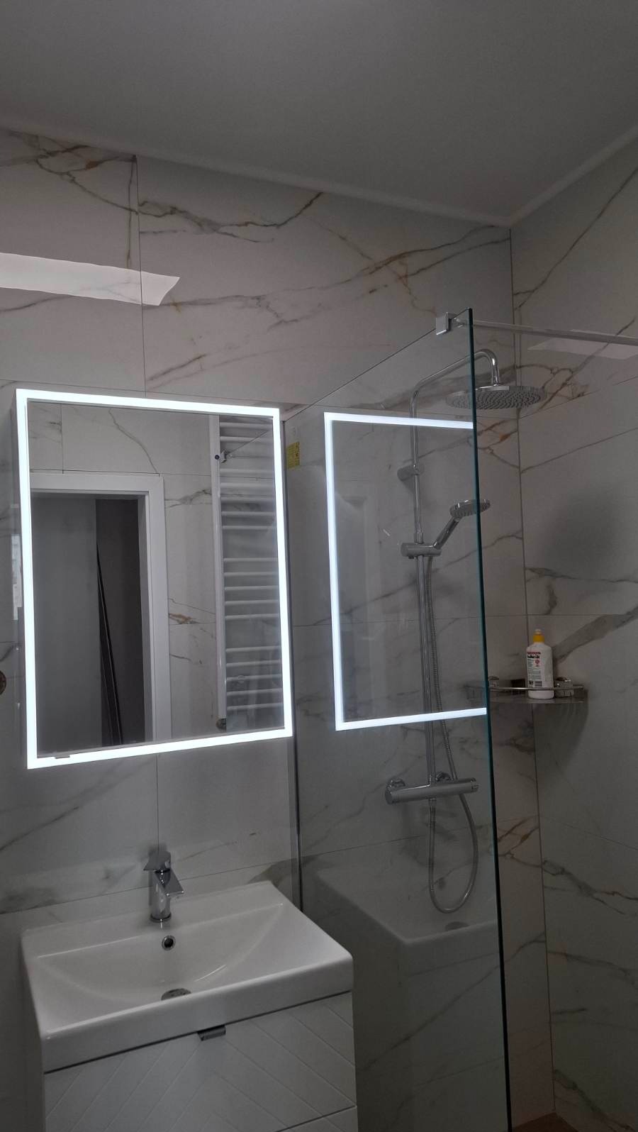 Nowoczesna łazienka z marmurowymi płytkami, podświetlanym lustrem i kabiną prysznicową ze szklaną ścianką. Widoczna umywalka z szafką i chromowana armatura.