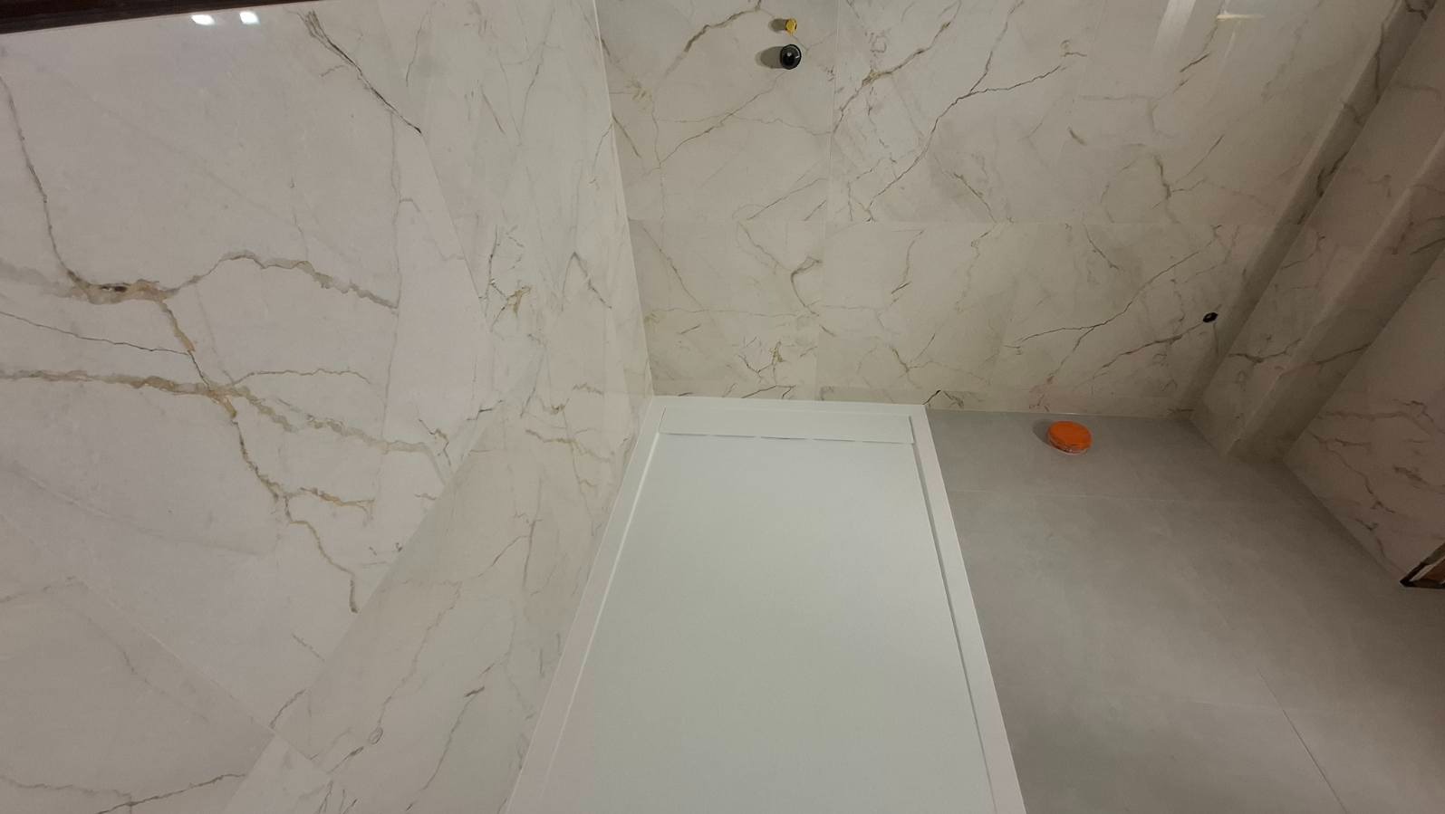 Wykończona kabina prysznicowa z białymi płytkami imitującymi marmur na ścianach i suficie. Widoczny odpływ liniowy i czujnik dymu.