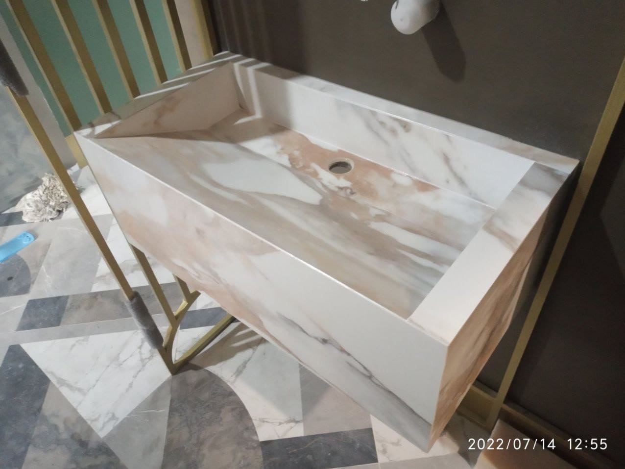 Marmurowa umywalka nablatowa na złotym stelażu, nowoczesny design. Umywalka z otworem na baterię, widoczna na tle szarej ściany i wzorzystej podłogi.