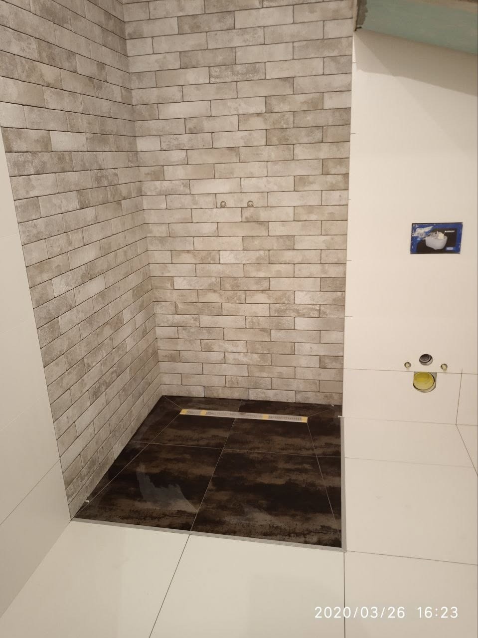 Wykończona strefa prysznicowa z beżową cegłą na ścianach i ciemnobrązową podłogą z odpływem liniowym. Białe płytki na pozostałej części podłogi i ścianie.