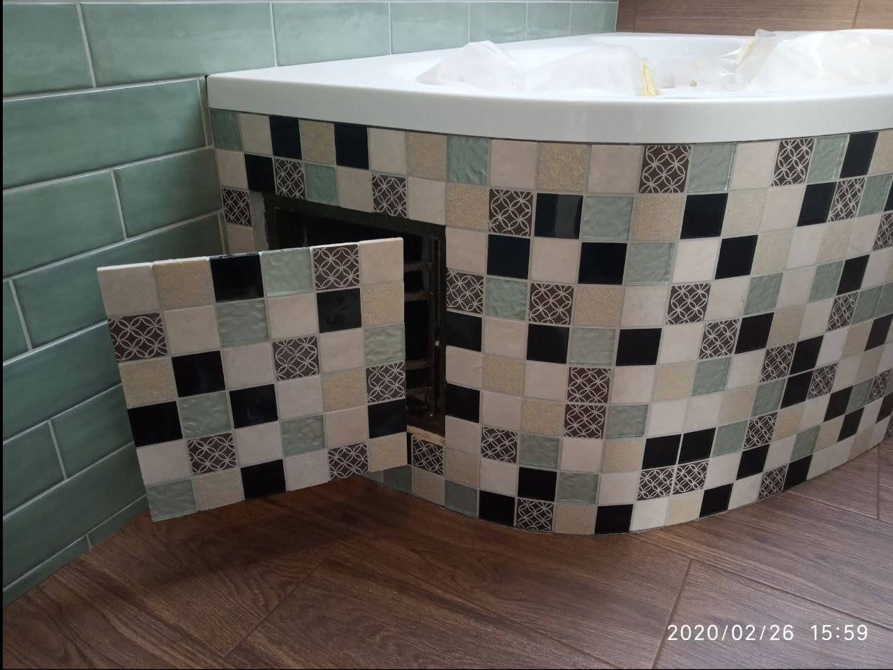 Obudowa wanny z mozaiki kwadratowej, z otwieraną klapką rewizyjną. Widoczne płytki w odcieniach beżu, czerni i zieleni. Podłoga wyłożona panelami imitującymi drewno.