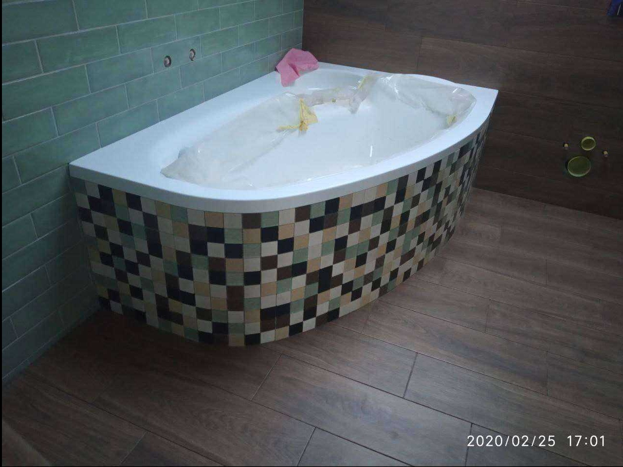 Narożna wanna obudowana mozaiką w odcieniach brązu i beżu, zabezpieczona folią. Na ścianie zielone płytki, na podłodze panele imitujące drewno. Widok z góry.