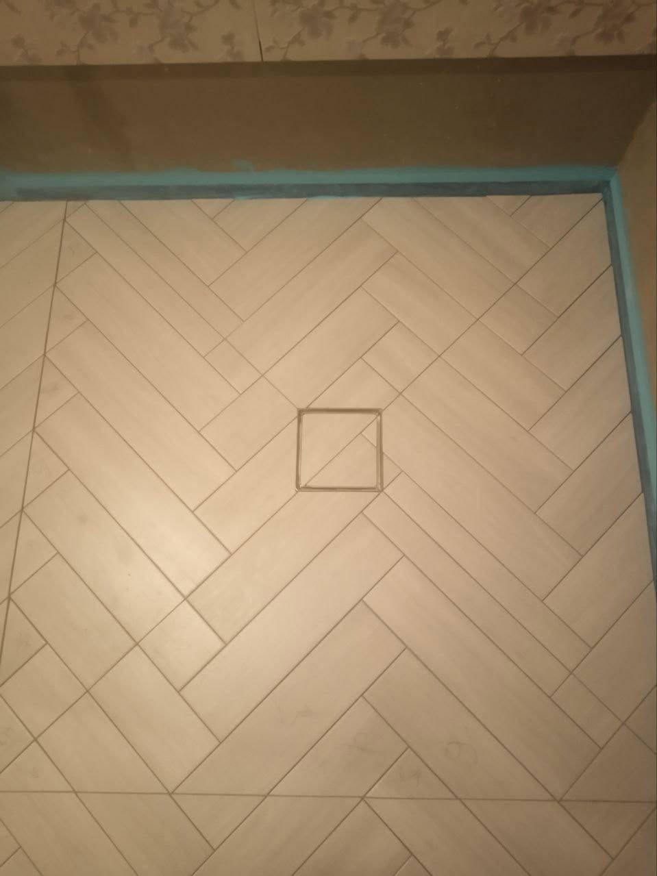 Beżowe płytki ułożone w jodełkę na podłodze prysznica z odpływem liniowym. Widoczne fugi i fragment ściany z tapetą.