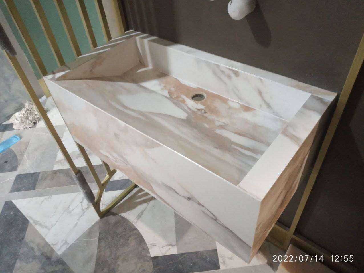 Marmurowa umywalka na złotym stelażu, nowoczesny design. Umywalka o geometrycznym kształcie, z widocznym wzorem marmuru. Całość w jasnej tonacji, na tle szarej ściany.