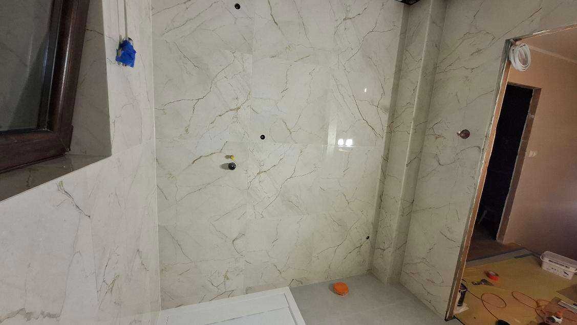 Wykończona łazienka z marmurowymi płytkami, widoczne przyłącza wodne i fragment okna. Na podłodze zabezpieczenia. Ujęcie z perspektywy podłogi.