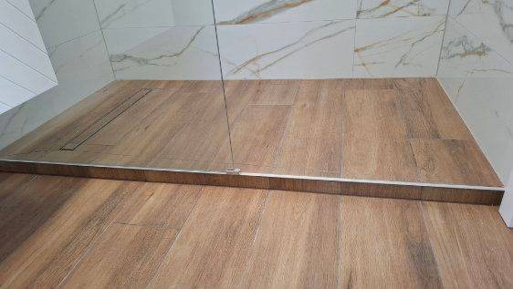 Nowoczesna kabina prysznicowa bez brodzika z odpływem liniowym, wykończona płytkami imitującymi drewno i marmur. Minimalistyczny design, jasne kolory.