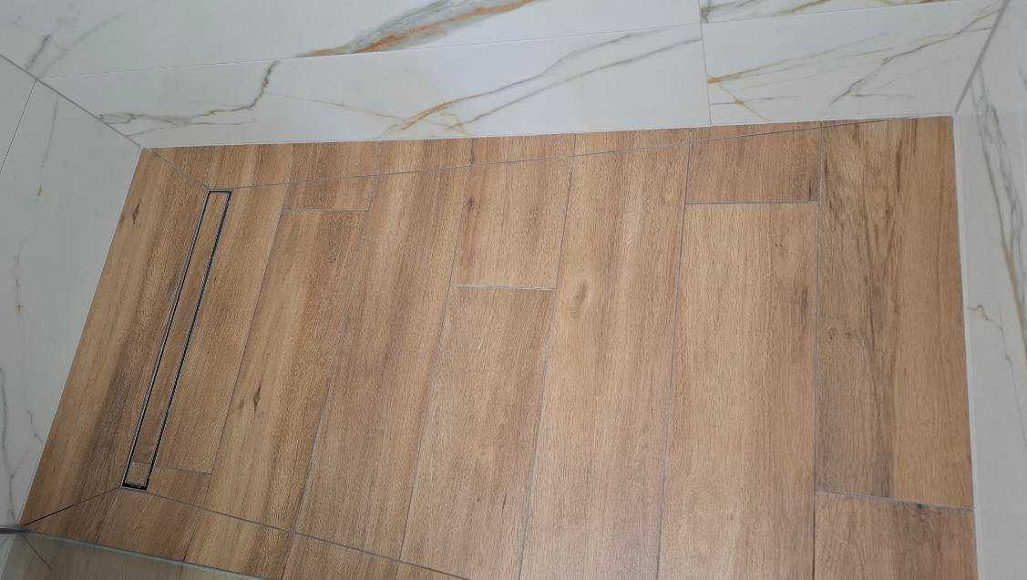 Wykończona podłoga w łazience z drewnopodobnych płytek, z odpływem liniowym i marmurowymi płytkami na ścianach. Detal wykończenia.