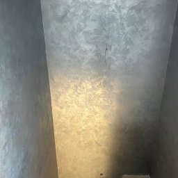 Zakład Remontowo Budowlany ART-DOM Dariusz Sajnóg - Wnętrze klatki schodowej z dekoracyjną szarą ścianą o nieregularnej fakturze, widoczne fragmenty schodów. Minimalistyczne wykończenie, nowoczesny design.