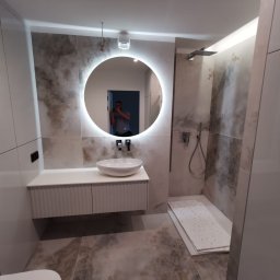 Zakład Remontowo Budowlany ART-DOM Dariusz Sajnóg - Elegancka łazienka z okrągłym lustrem LED, umywalką nablatową i kabiną prysznicową typu walk-in. Płytki imitujące marmur i białe meble tworzą nowoczesny design.