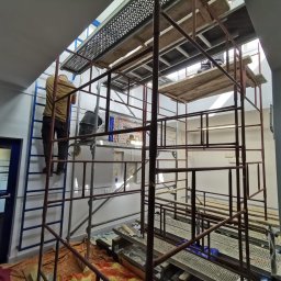 Zakład Remontowo Budowlany ART-DOM Dariusz Sajnóg - Prace remontowe w biurze: pracownik na rusztowaniu, szpachlowanie sufitu. Widoczne narzędzia i materiały budowlane. Charakterystyczne oświetlenie w tle.