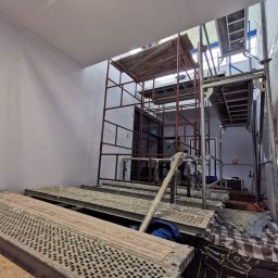 Zakład Remontowo Budowlany ART-DOM Dariusz Sajnóg - Wnętrze w trakcie malowania: białe ściany, rusztowania metalowe, widoczny pracownik. Przygotowanie do malowania w wysokim pomieszczeniu z oknem dachowym.