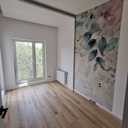 Zakład Remontowo Budowlany ART-DOM Dariusz Sajnóg - Jasne panele podłogowe w pustym pokoju z oknem i dekoracyjną tapetą z motywem kwiatowym na ścianie. Widoczny grzejnik i wystające kable ze ściany.
