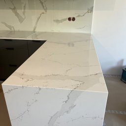 Zakład Remontowo Budowlany ART-DOM Dariusz Sajnóg - Biały blat kuchenny z marmurowym wzorem, dopasowany do płytek na ścianie, widok na czarne szafki i gniazdka elektryczne, ukazujący estetyczne wykończenie.