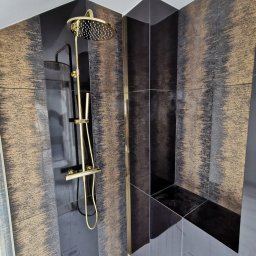 Zakład Remontowo Budowlany ART-DOM Dariusz Sajnóg - Wykończona kabina prysznicowa z czarno-złotymi płytkami i złotą armaturą, w nowoczesnym stylu. Widoczne półki w zabudowie i eleganckie wykończenie.