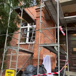 Zakład Remontowo Budowlany ART-DOM Dariusz Sajnóg - Ceglana elewacja budynku w trakcie renowacji, osłonięta rusztowaniem z tabliczką ostrzegawczą 'Roboty na wysokości', zabezpieczona taśmą. Widoczne okno i wejście.
