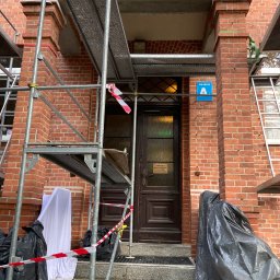 Zakład Remontowo Budowlany ART-DOM Dariusz Sajnóg - Renowacja elewacji budynku z czerwonej cegły. Widoczne rusztowania, zabezpieczenia i drzwi wejściowe z tabliczką informacyjną. Prace w toku.