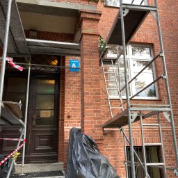 Zakład Remontowo Budowlany ART-DOM Dariusz Sajnóg - Ceglana fasada budynku w trakcie renowacji, z widocznymi rusztowaniami, wejściem i oknami. Część elewacji zakryta folią ochronną. Tabliczka informacyjna przy wejściu.