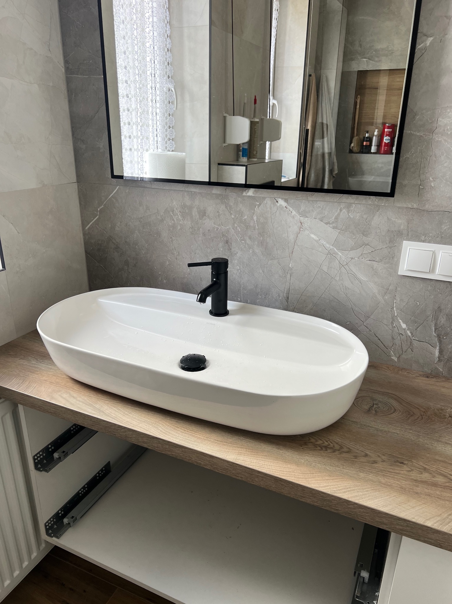 Nowoczesna, biała umywalka nablatowa z czarną armaturą na drewnianym blacie w łazience wykończonej szarą marmurową płytką. W tle lustro i akcesoria.