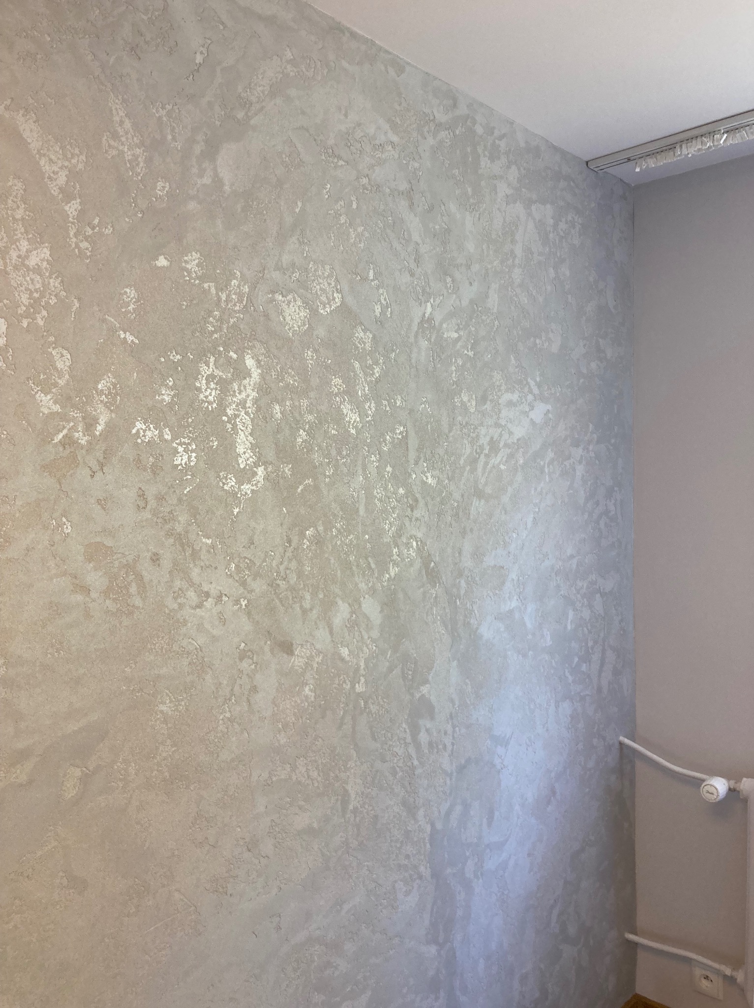 Ściana z dekoracyjnym, srebrzystym tynkiem strukturalnym w rogu pomieszczenia. Widoczny fragment grzejnika i karnisz.