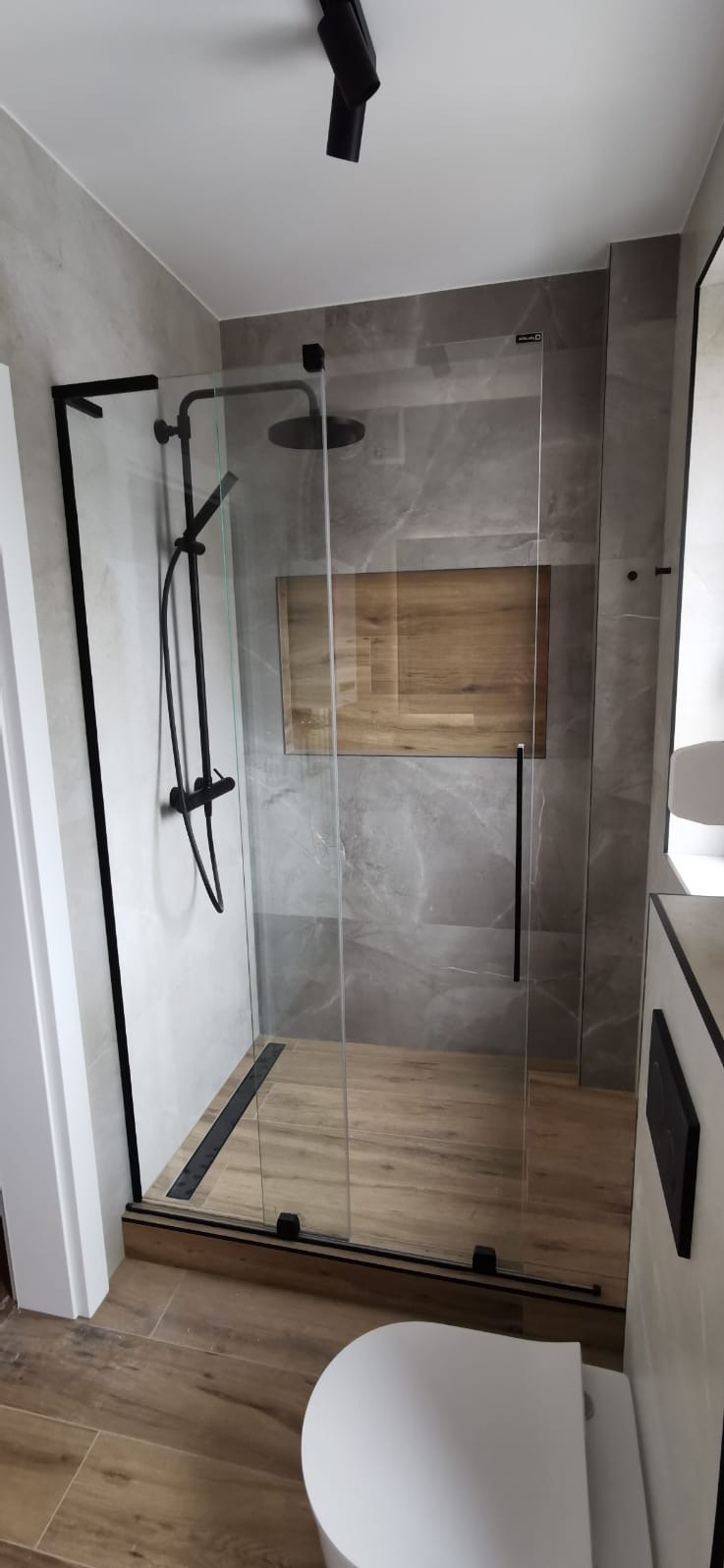 Nowoczesna kabina prysznicowa walk-in z czarnymi detalami i drewnopodobną podłogą, w łazience wykończonej szarymi płytkami imitującymi marmur.