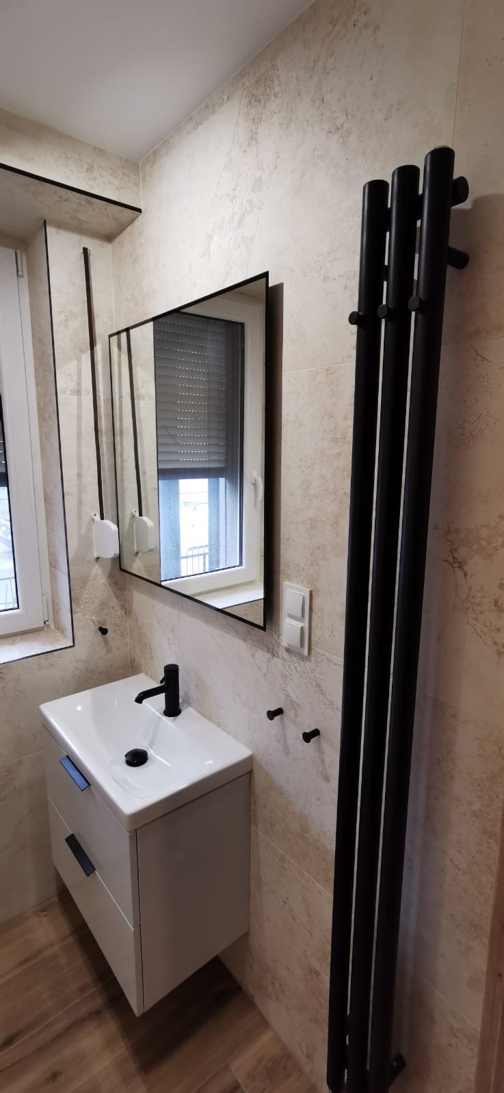 Nowoczesna łazienka z beżowymi płytkami: biała szafka pod umywalką z czarną armaturą, czarny grzejnik drabinkowy i lustro z czarną ramą odbijające okno z żaluzjami.