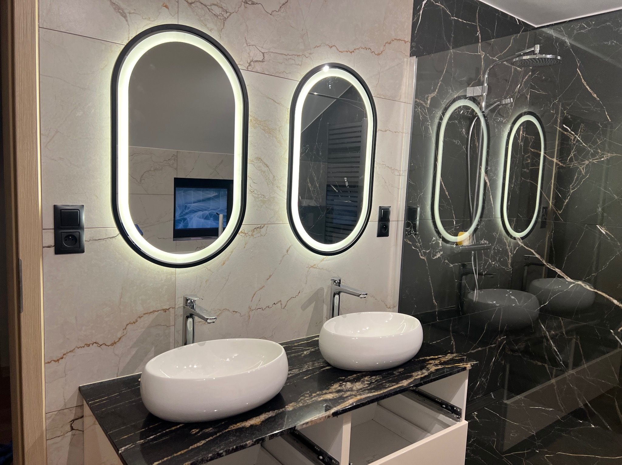 Nowoczesna łazienka z dwoma umywalkami nablatowymi, podświetlanymi lustrami owalnymi i marmurowymi płytkami w odcieniach bieli i czerni.