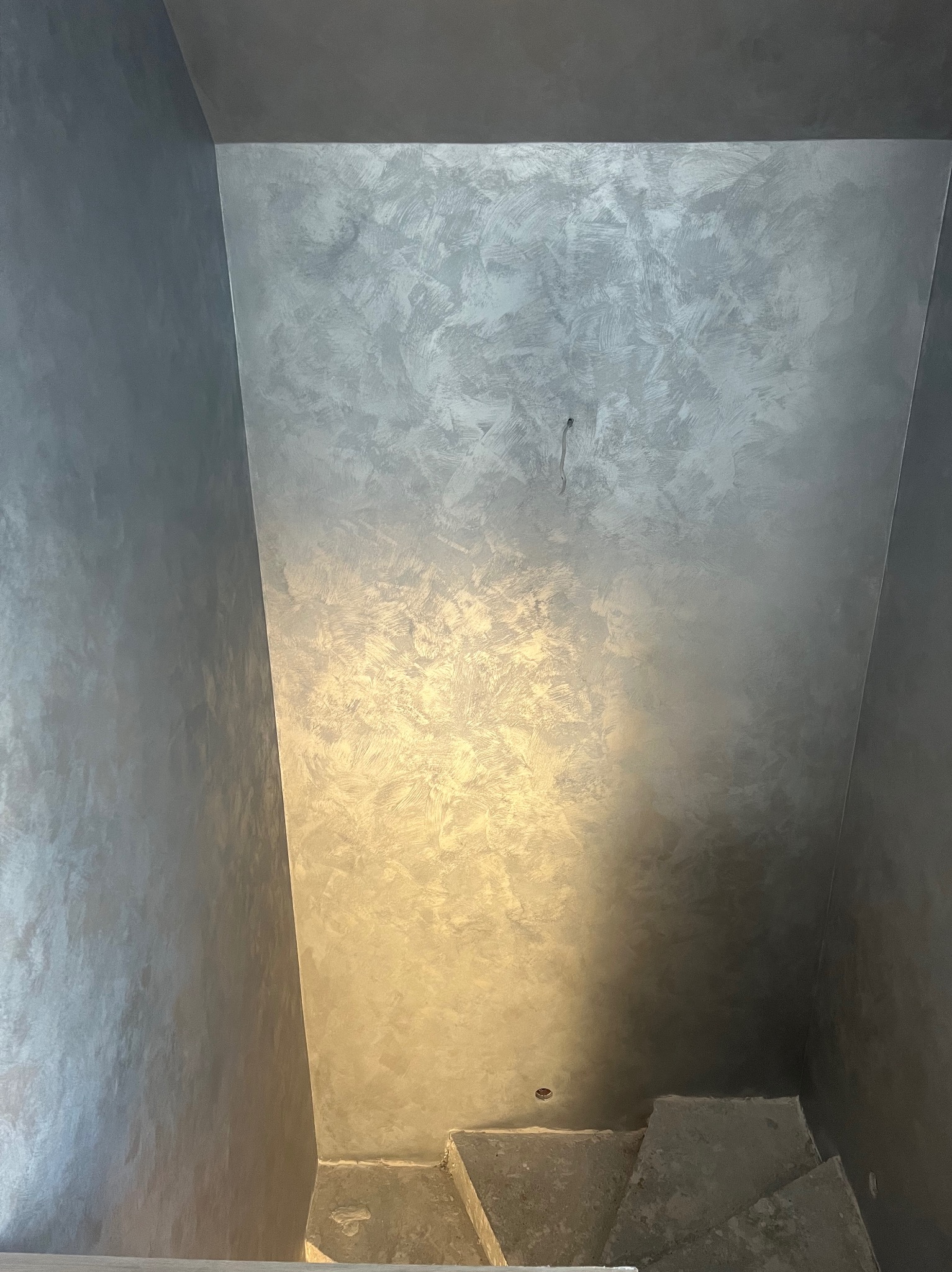 Wnętrze klatki schodowej z dekoracyjną szarą ścianą o nieregularnej fakturze, widoczne fragmenty schodów. Minimalistyczne wykończenie, nowoczesny design.