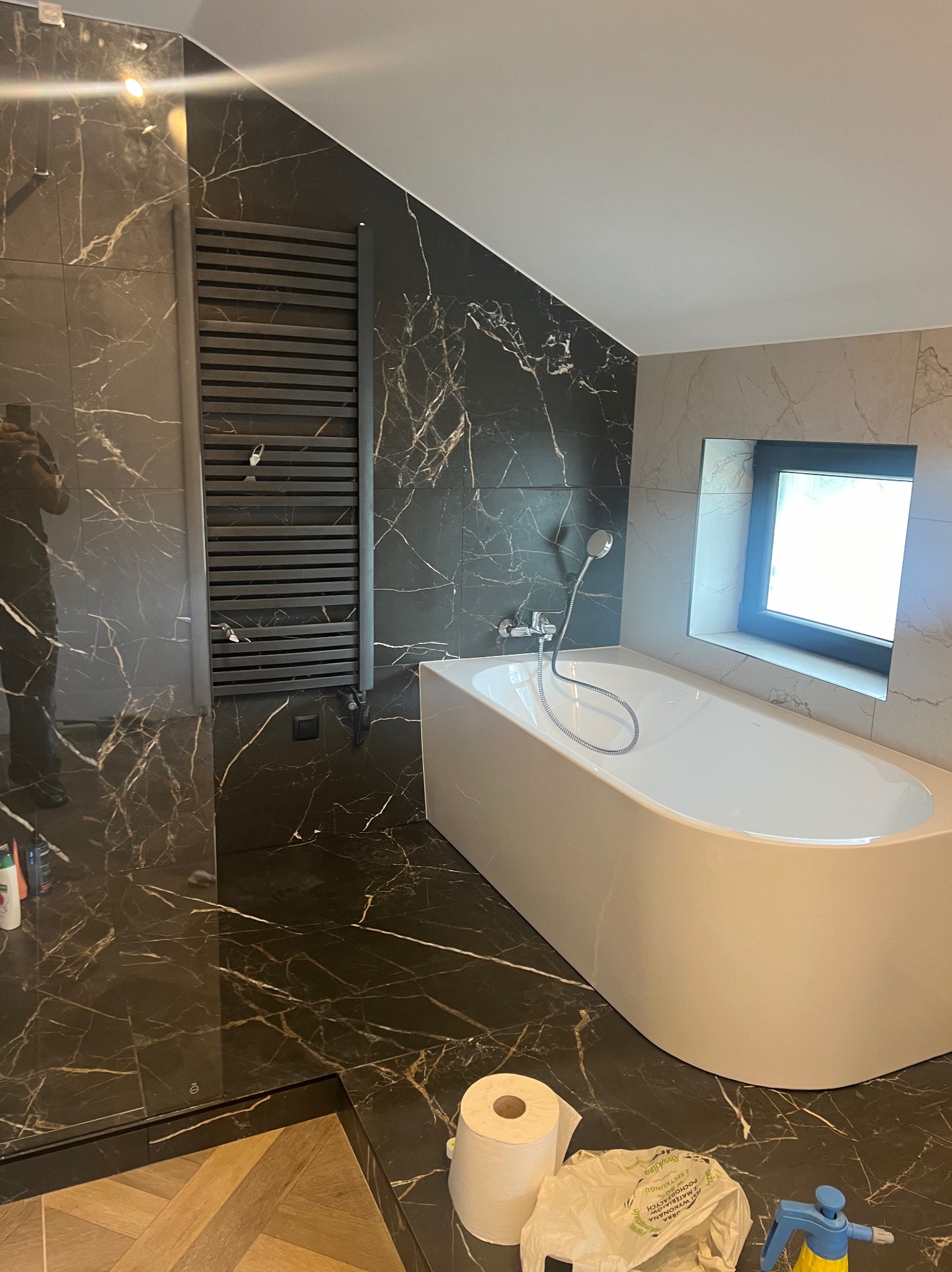 Nowoczesna łazienka z czarnymi płytkami imitującymi marmur, białą wanną i grzejnikiem drabinkowym. Okno z czarną ramą doświetla wnętrze.