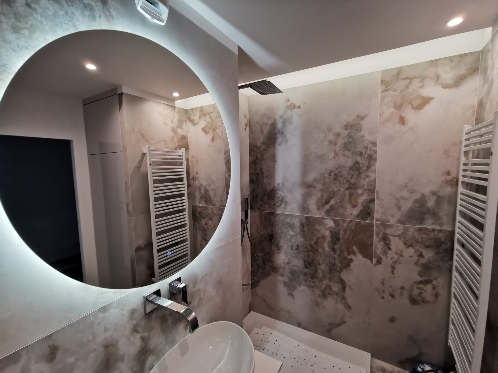 Wykończona łazienka z okrągłym lustrem z podświetleniem LED, umywalką nablatową i grzejnikiem drabinkowym na ścianie w neutralnych, marmurkowych płytkach.