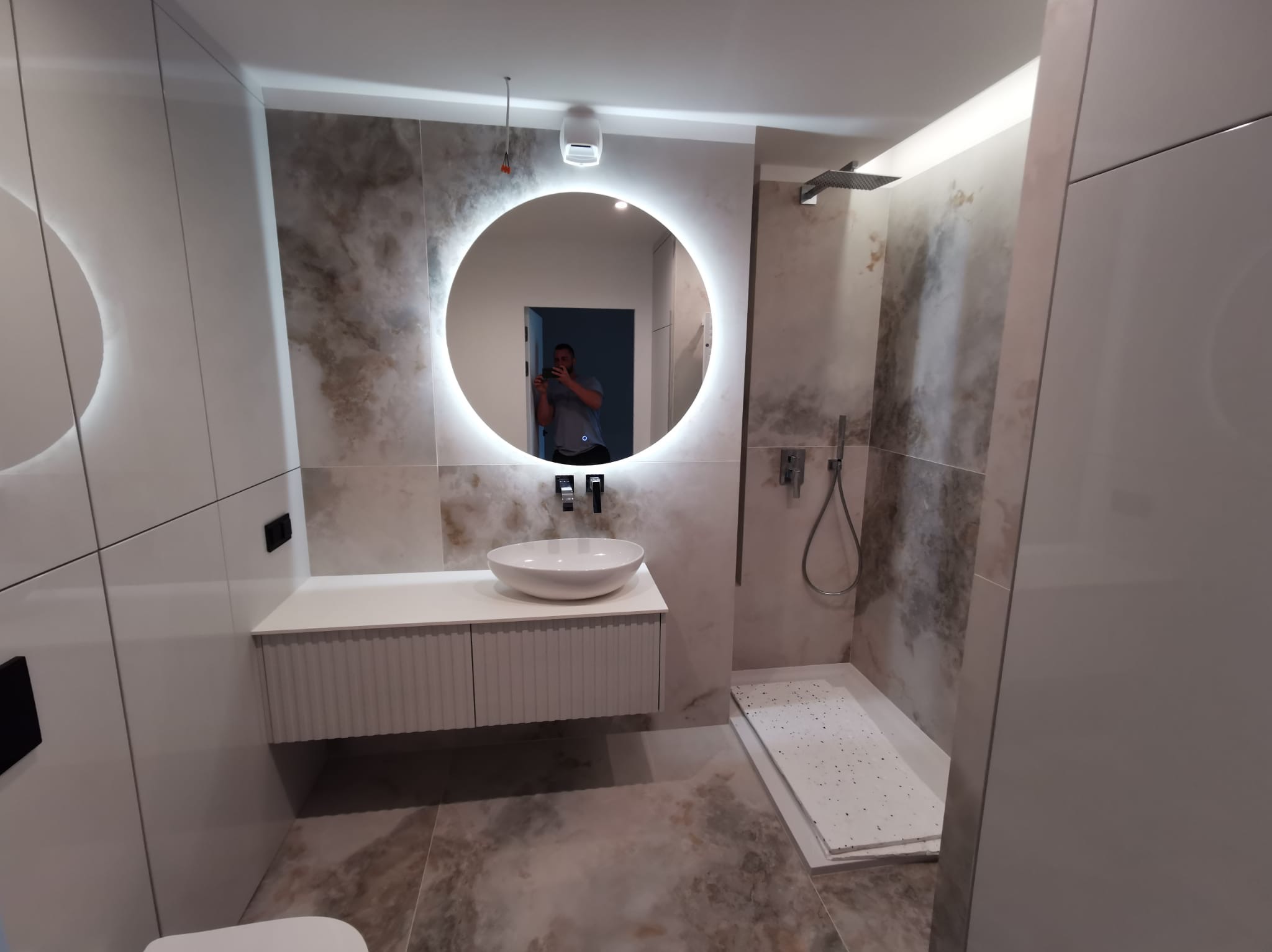 Elegancka łazienka z okrągłym lustrem LED, umywalką nablatową i kabiną prysznicową typu walk-in. Płytki imitujące marmur i białe meble tworzą nowoczesny design.