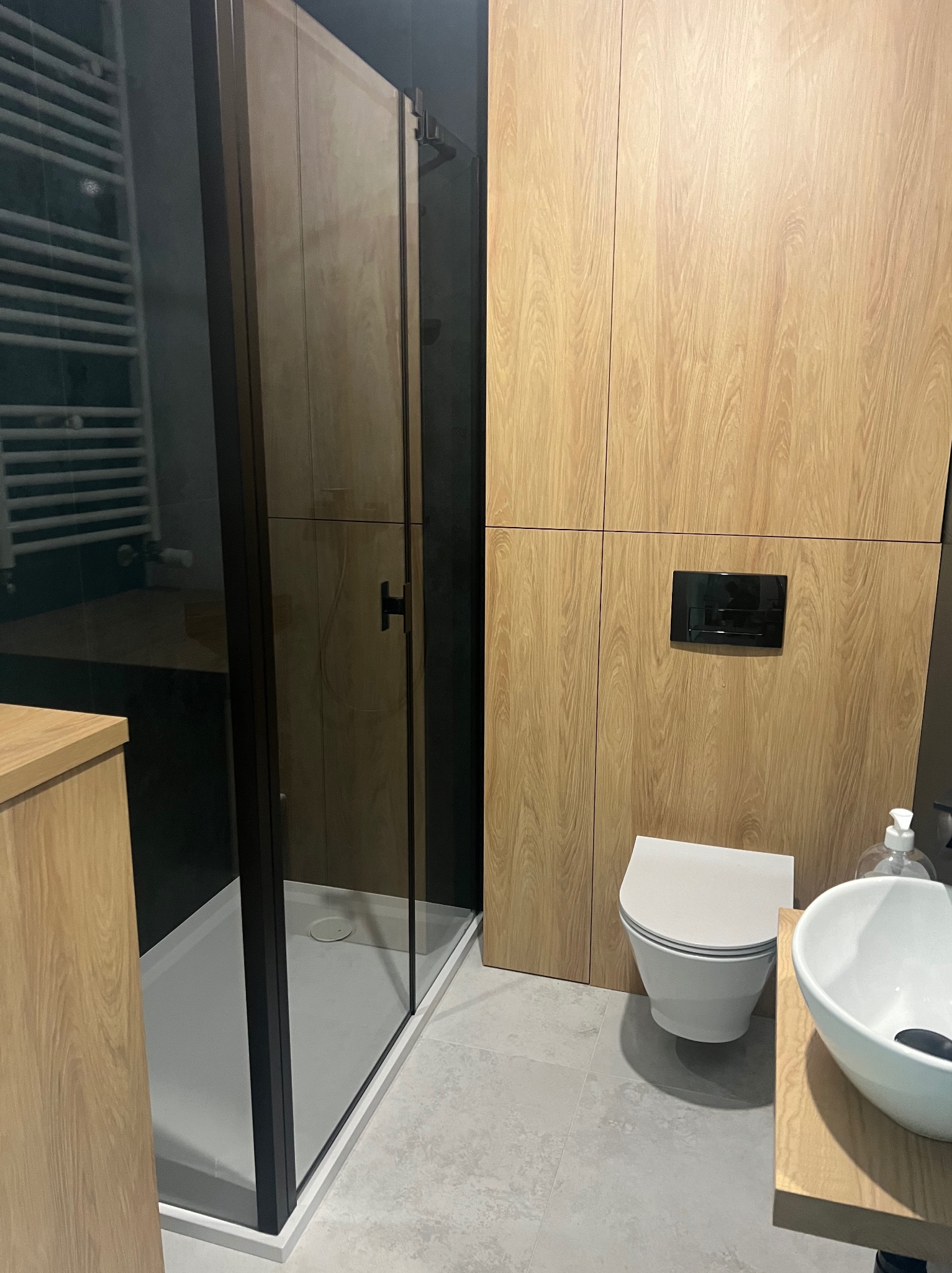 Nowoczesna łazienka z kabiną prysznicową typu walk-in, drewnianymi szafkami i umywalką nablatową. Minimalistyczny design z czarnymi akcentami.