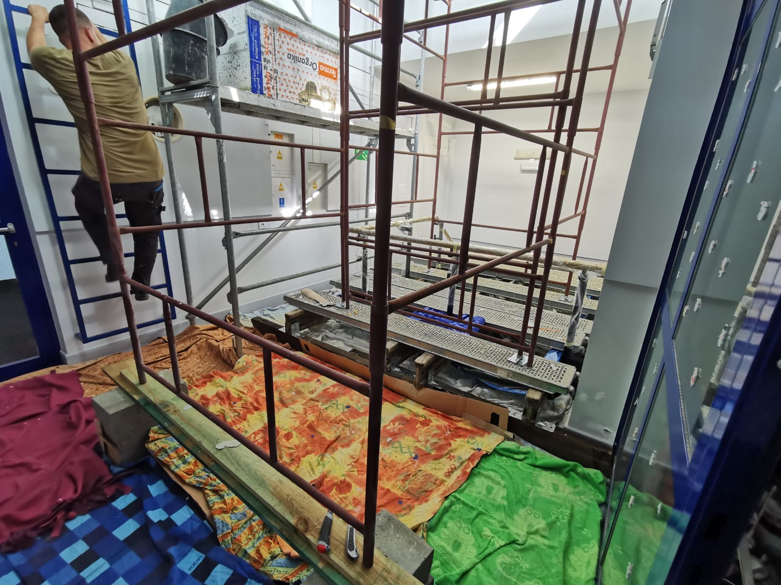 Wnętrze w trakcie prac wykończeniowych: rusztowanie, zabezpieczona podłoga, pracownik wspinający się po drabinie. Widoczna puszka produktu termoizolacyjnego.