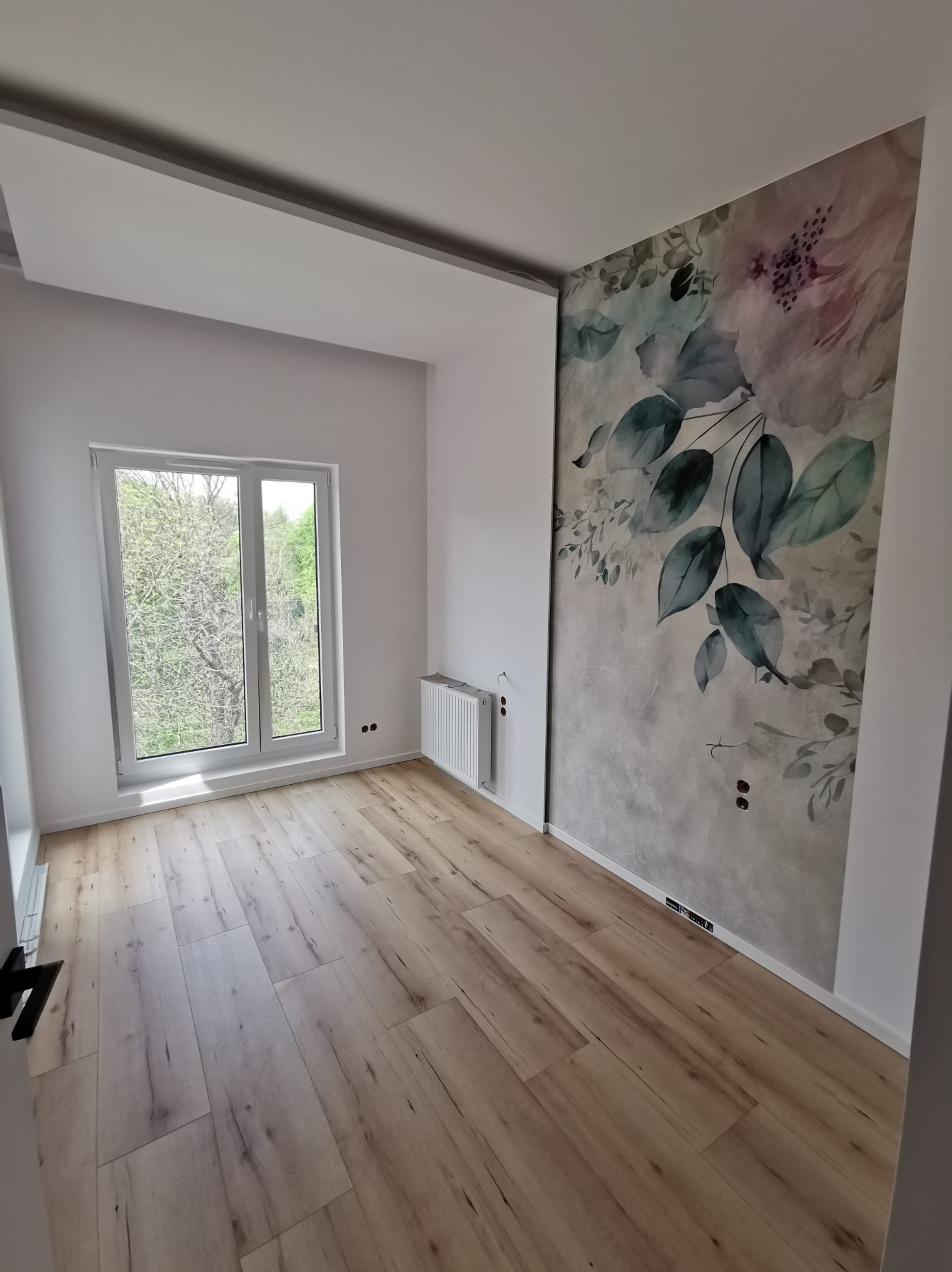 Jasne panele podłogowe w pustym pokoju z oknem i dekoracyjną tapetą z motywem kwiatowym na ścianie. Widoczny grzejnik i wystające kable ze ściany.