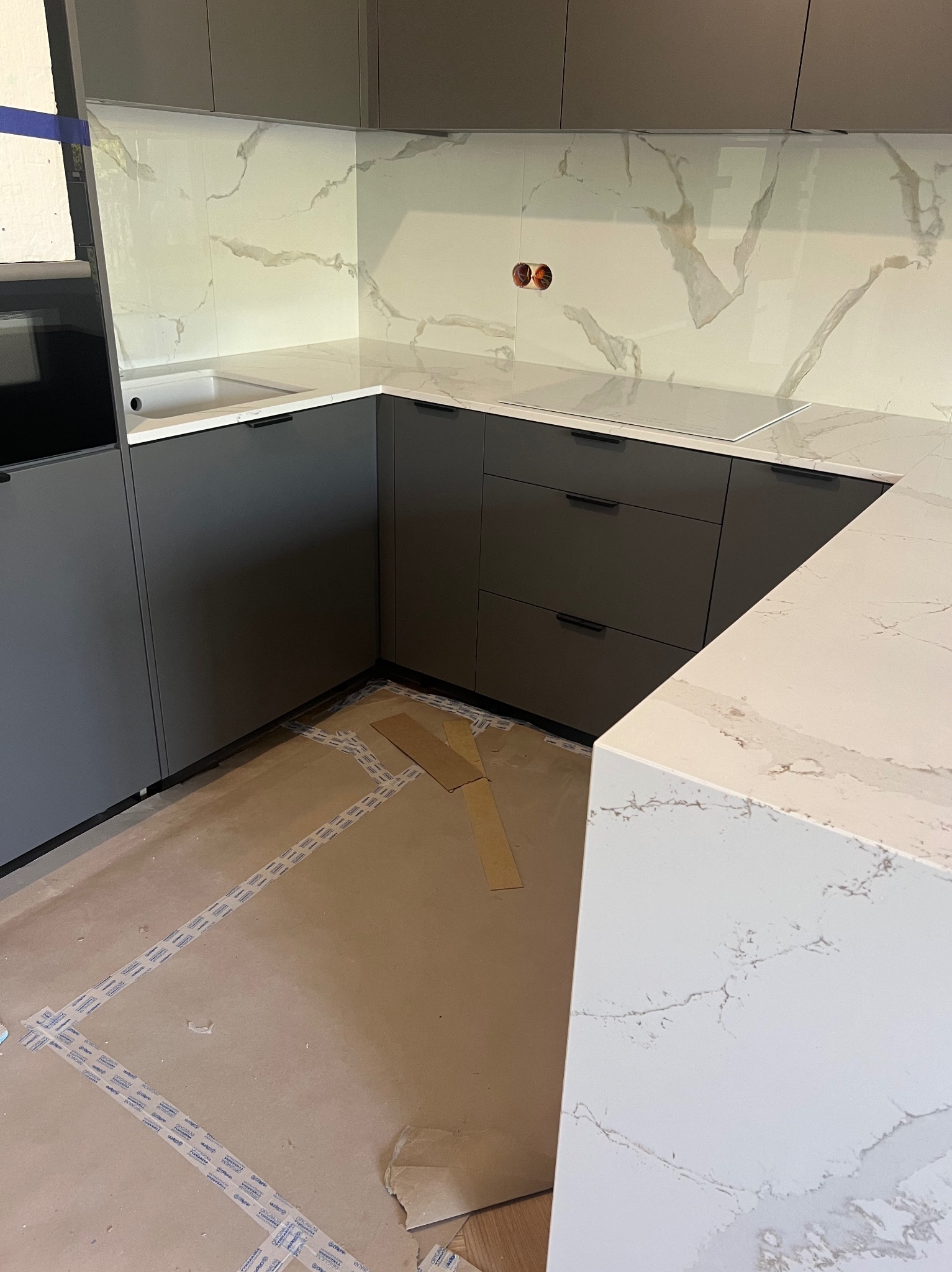 Nowoczesna, szara zabudowa kuchenna z blatem imitującym marmur i szklanym panelem ściennym z motywem marmuru. Podłoga zabezpieczona kartonem.